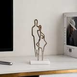 Decorative sculpture of two figures on a marble base on a shelf with a magazine in the background.