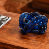 A translucent dark blue glass knot sculpture resting on a polished wooden table, with a blurred magazine partially visible on the left.