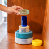 A hand is placing a small textured green glass ring onto a stack of stacked glass vessels in blue, cream, teal, white, and yellow, set against a warm wooden background.