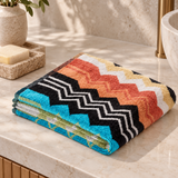 A folded multicolor chevron-patterned towel sits on a beige marble bathroom counter next to a soap dish with a bar of soap and a potted plant.