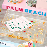 Clear decorative box with gold glitter and a small white card with green designs, on a surface with books in the background.