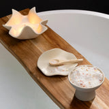 Wooden tray with a bowl of bath salts, a spoon, and two shell-shaped candle holders on a bathtub.