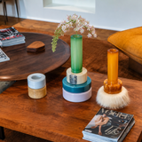 A styled interior scene with a wooden coffee table holding decorative objects, including a green glass vase with white flowers, stacked round containers, and a translucent amber candle holder with a white faux-fur base. A mustard yellow armchair and another round wooden table are visible in the room.