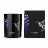 A black scented candle jar with a hummingbird and purple flowers printed on the side, shown next to its matching black box featuring the same artwork. The brand name Vila Hermanos is visible on both the candle and the box.
