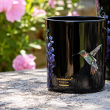 Two black ceramic candle jars with hummingbird and wisteria floral designs, displayed on a stone surface with a blurred garden background of pink flowers.
