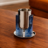 A polished silver-toned metal cup holder with a matching saucer, featuring a central cylindrical insert surrounded by four blue marbled square blocks, displayed on a wooden table.
