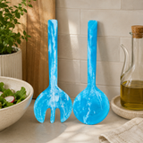 Two blue salad servers on a kitchen counter with a bowl of salad and oil bottle in the background.
