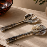 Two decorative spoons on a textured surface with a bowl and vase in the background.