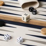 A close-up of a backgammon board with cream and navy striped playing surface, wooden double-six dice, black and white checkers, and a cream canvas bag.
