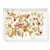A clear acrylic tray filled with scattered gold foil flakes and assorted colorful capsules and pills, photographed from above on a white background.