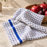 A white kitchen towel with a blue grid pattern and a solid blue stripe, draped over a marble countertop next to a wooden bowl of pomegranates.